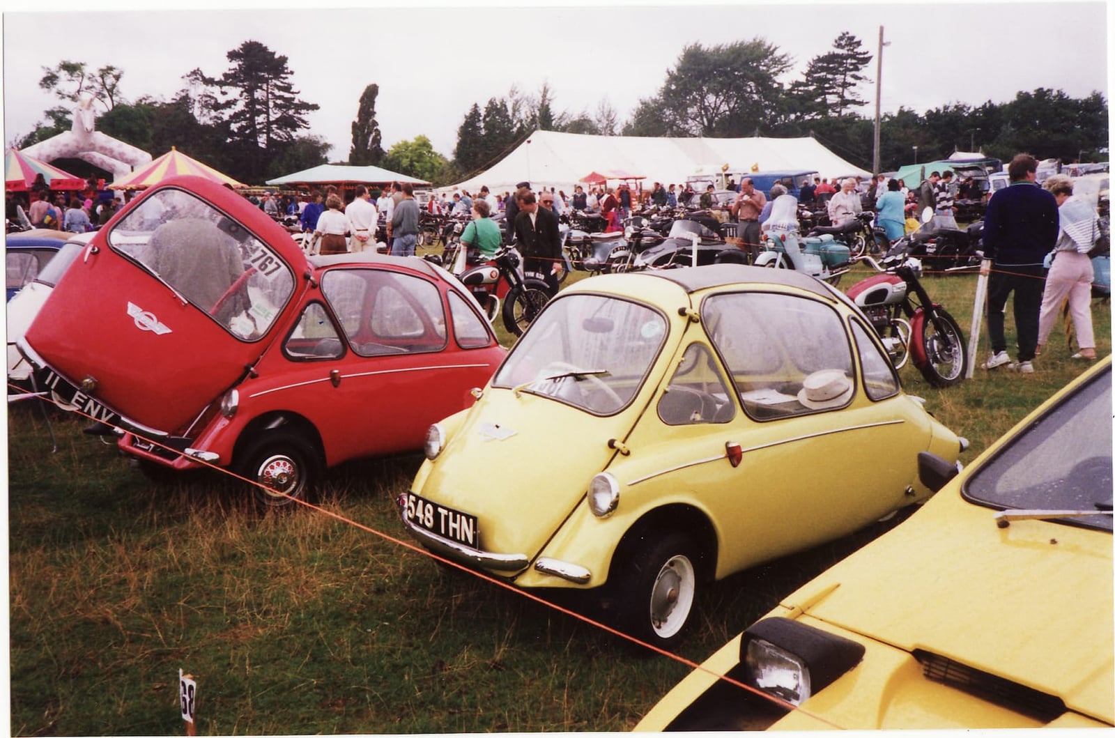 Two Trojan 200 bubble cars, one red and one yellow, on grass at a British show with crowds behind