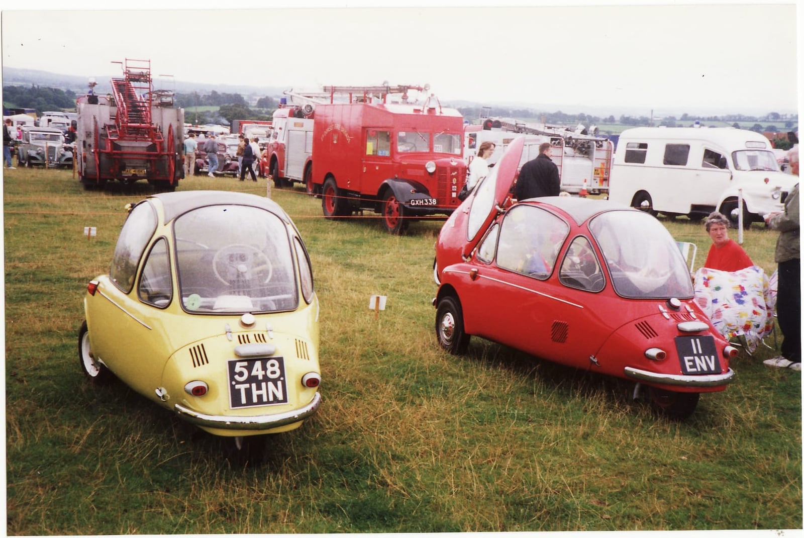 Two Trojan-built Heinkel bubble cars, one cream and one red, on grass at a classic vehicle rally