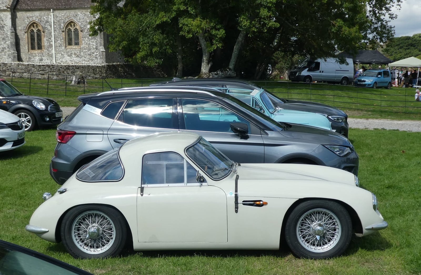 A white 1959 TVR Grantura in side profile at a classic car show beside a church