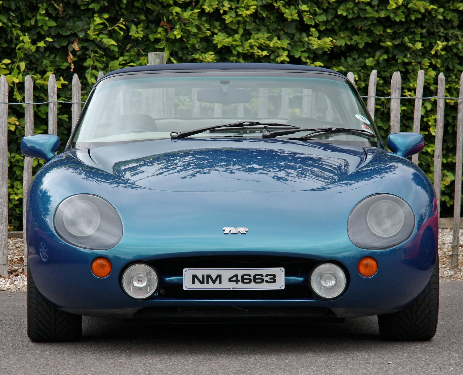 A metallic blue-green TVR Griffith convertible, head-on front view, against a wooden fence and hedge